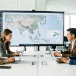 Équipe technologique internationale composée de deux femmes et deux hommes analysant une carte sans texte de l’Asie du Sud-Est sur de grands écrans dans un bureau moderne et lumineux.